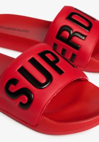 Røde slidesandaler lavet af syntetisk materiale med en bred rem med hævet sort "SUPER" skrift. Tekstureret sål for bedre greb.