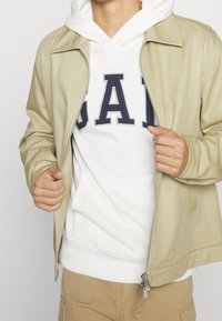 Giacca beige con zip sopra un hoodie bianco caratterizzato da lettere in blu marine. La giacca ha una texture liscia e un collare standard, con tasche frontali.