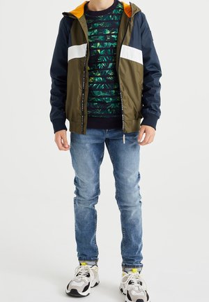 Veste légère à blocs de couleurs en vert olive et bleu marine avec des rayures blanches, associée à un pull à motifs et un jean bleu. Chaussures de sport incluses.