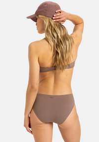 Braunfarbener Bikini aus strukturiertem Stoff, mit einem Bandeau-Oberteil und einem leicht hochgeschnittenen Unterteil. Eine passende braune Mütze wird getragen, das Haar fällt über den Rücken.