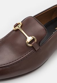 Καφέ δερμάτινα loafers με λεπτομέρεια από χρυσό τόνο, διαθέτοντας στρογγυλή μύτη και λεία, γυαλισμένη επιφάνεια.