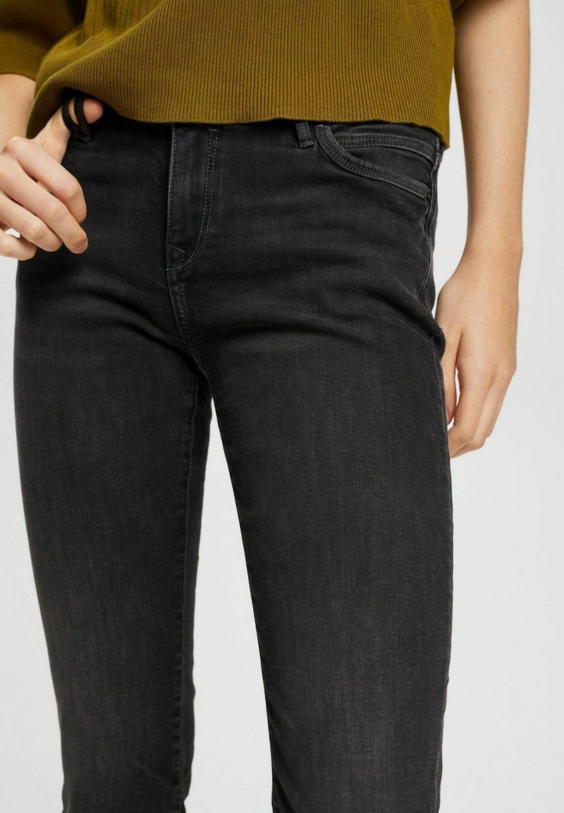 Jean en denim noir à coupe slim, avec une taille mi-haute, des coutures discrètes et une poche frontale. Le tissu a une texture lisse.