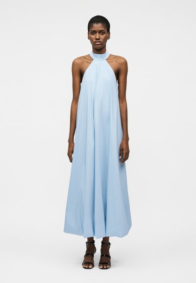 Femme aux cheveux courts portant une robe maxi sans manches bleu clair à col licou et des talons noirs ouverts, debout devant un fond blanc uni.
