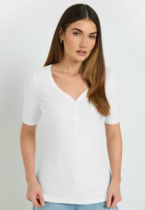 Woman with long brown hair wears a white short-sleeve ribbed shirt with three buttons, standing against a plain light background.