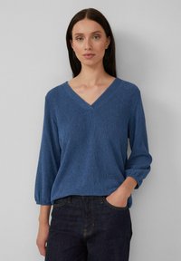 Femme portant un blouse bleu texturé à col en V avec des manches trois-quarts et un jean en denim foncé, se tenant devant un fond gris uni.