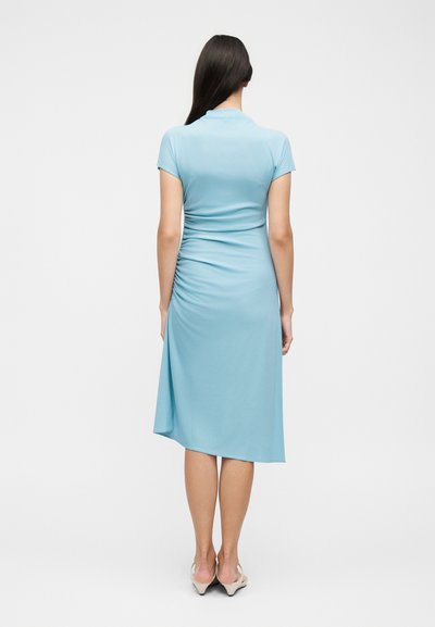 Γυναίκα με μακριά σκούρα μαλλιά όρθια, φοράει ένα ανοιχτό μπλε φόρεμα με κοντά μανίκια, μήκους μέχρι τη μέση της γάμπας, με λεπτομέρεια ρέλι και μπεζ χαμηλά τακούνια.