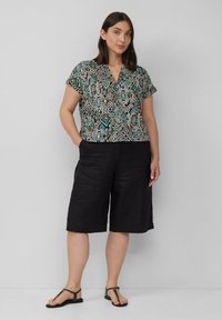 Multikleurige overhemden met korte mouwen met geometrische patronen, gecombineerd met zwarte linnen shorts en zwarte platte sandalen. Neutrale achtergrond.