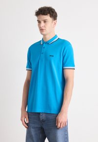Plava polo majica s kratkim rukavima, tri gumba i kontrastnim obrubom ovratnika. Sadrži crni logo "BOSS" na lijevoj strani prsa.