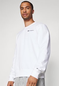 Sweatshirt en polaire blanche à manches longues, col rond et logo brodé sur la poitrine. Présente des poignets et un ourlet côtelés.