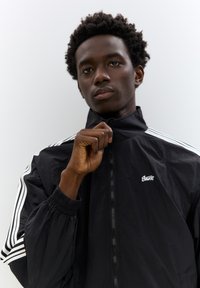 Sort zip-up jakke med hvide striber på ærmerne, der har en høj krave og elastiske manchetter; letvægts, glat materiale, lille logo.