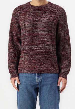 Mężczyzna ubraný w bordowy i szary sweter z teksturowanym splotem, z długimi rękawami, oraz niebieskie dżinsy, stoi na tle jednolitego tła.