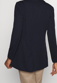 Blazer bleu marine à manches longues avec finition texturée, coupe ajustée et ourlet légèrement incurvé. Comprend une fente dorsale unique et un col cranté.