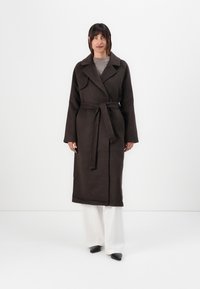 Long brown coat with a wrap design, wide lapels, belted waist, and long sleeves. Worn over a light gray top and white trousers.