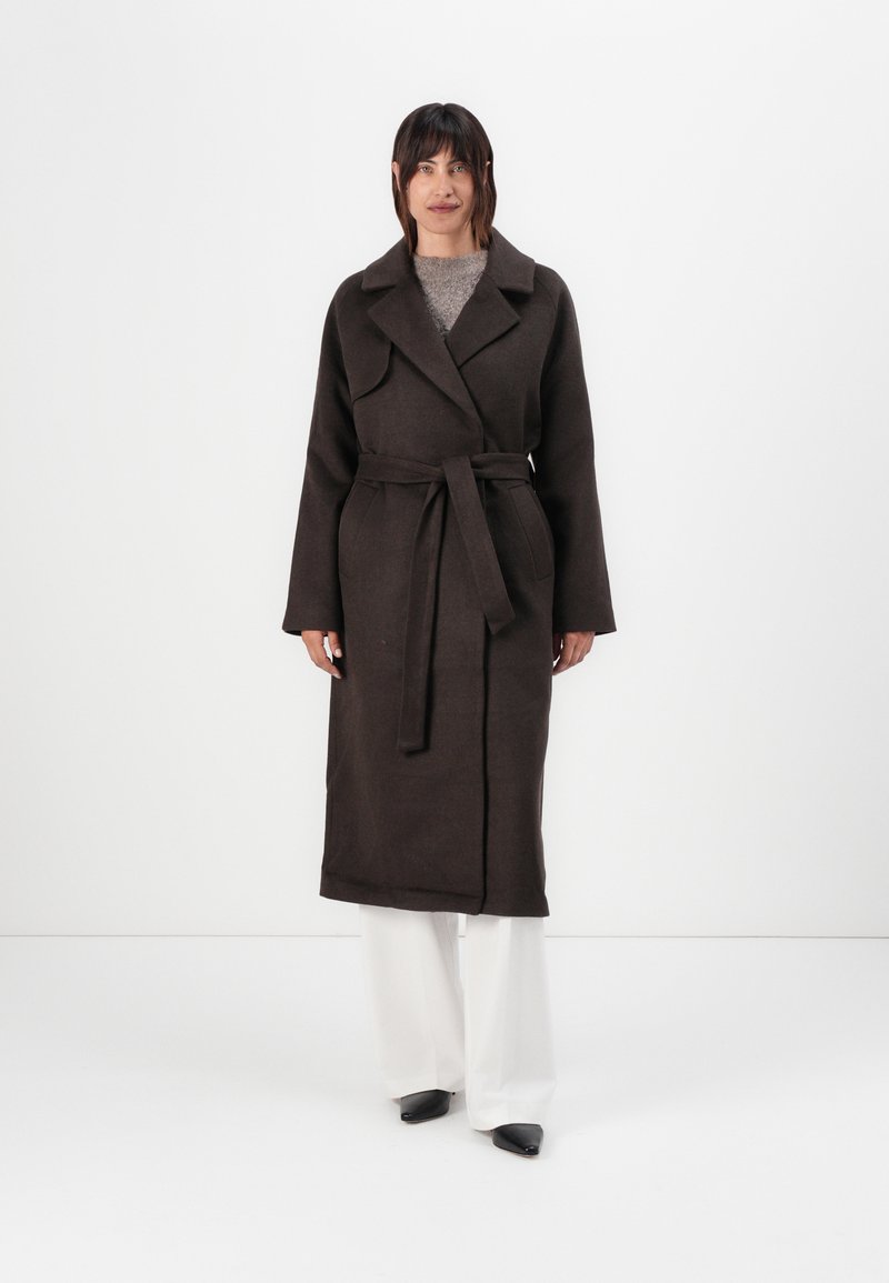 Long brown coat with a wrap design, wide lapels, belted waist, and long sleeves. Worn over a light gray top and white trousers.