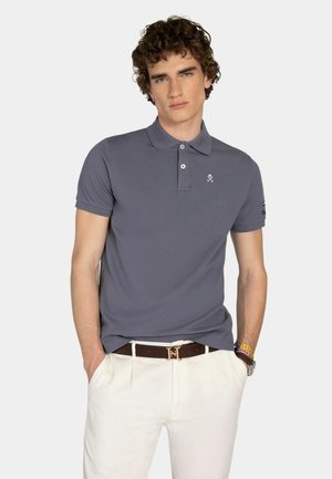 Hombre con cabello rizado, vistiendo una camisa polo gris y pantalones blancos con un cinturón marrón, con las manos en los bolsillos, de pie frente a un fondo liso.