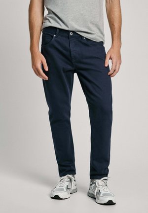 Chinos azul marino hechos de algodón con una textura suave, que presentan un diseño clásico de cinco bolsillos y un cierre de botón. Llevados con zapatillas grises.