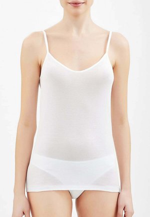 Donna che indossa una canottiera bianca aderente e biancheria intima bianca, in piedi davanti a uno sfondo bianco uniforme, rivolta in avanti.