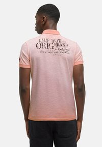 Poloshirt in lichtroze met een vervaagd ontwerp; heeft zwarte grafische tekst op de achterkant. Korte mouwen met een contrasterende kraag. Katoenen materiaal.