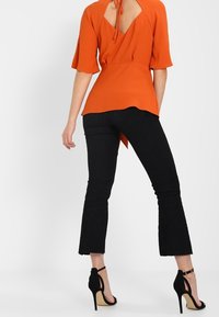 Blouse orange à manches courtes avec un lien à nouer dans le dos, associée à un pantalon noir évasé et des sandales noires à talons hauts. Tissu lisse, lignes épurées.