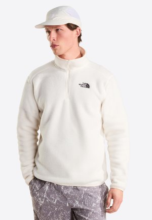 Ung mand iført hvid fleecejakke med kvart-lynlås og The North Face-logo, hvid mesh-kasket og grå mønstrede bukser, vendt mod venstre.