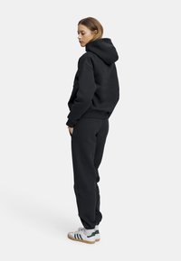 Svart hettegenser med hette, avslappet passform, kombinert med matchende svarte tapered joggebukser. Hvite joggesko med grønne detaljer.