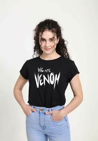 Černé tričko s krátkým rukávem s tlustým bílým nápisem "JSME VENOM." V kombinaci se světlomodrými džíny s vysokým pasem, což vytváří ležérní vzhled.