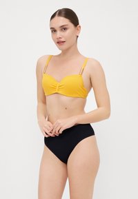 Gul bikini topp med rynkede detaljer og gyldne aksentstropper, parret med høytlivede svarte bikini-bukser. Glatt tekstur og minimalt design.