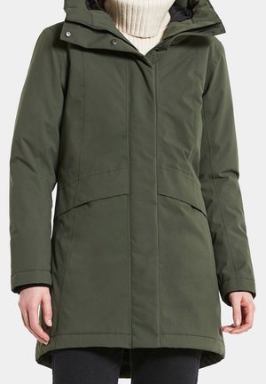 Femme portant un parka d’hiver vert olive avec un col montant sur un pull à col roulé côtelé crème et un pantalon noir.