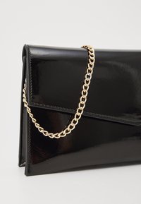 Pochette en cuir verni noir avec une bandoulière en chaîne dorée, présentant un design à rabat triangulaire et une finition douce et brillante.