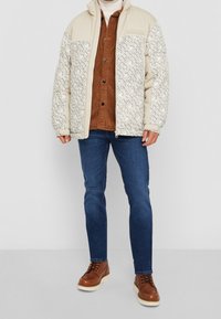 Kurtka puchowa w jasnym beżu z wzorzystym przodem; zestawiona z brązową koszulą z sztruksu i niebieskimi dżinsami; brązowe buty za sznurówki.