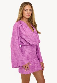 Robe rose courte avec de longues manches évasées présentant des motifs floraux texturés. Le décolleté en V et la taille cintrée créent une silhouette ajustée.