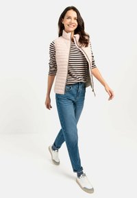 Lichtroze gewatteerde vest, met rits, gedragen over een zwart-wit gestreept shirt met lange mouwen. Blauwe jeans en witte sneakers maken de outfit compleet.