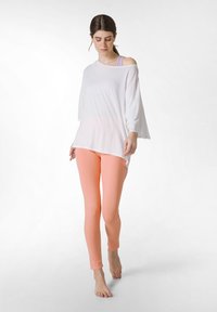 Witte, losse top met brede mouwen, gecombineerd met perzikkleurige leggings. Model staat op blote voeten en toont de ontspannen pasvorm van de outfit.