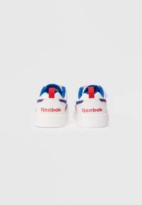 Bijele Reebok tenisice s crvenim logotipom na peti, plavim jastučićem unutar, i crvenom povučnom trakom, prikazane iz pozadine na jednostavnoj bijeloj pozadini.
