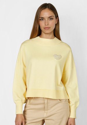 Woman wearing a pale yellow sweater with a pearl heart decoration on the left chest, paired with light beige pants.