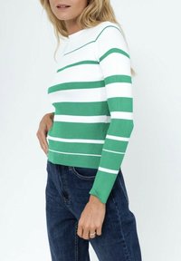 Pull en tricot rayé vert et blanc avec des manches longues, un col rond et un tissu texturé ; porté avec un jean bleu foncé.