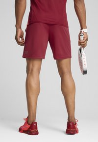 Tamsiai raudoni sportiniai šortai su tinklinio tekstūra, derinami su raudonais sportiniais bateliais, turinčiais baltų ir rožinių akcentų. Asmuo laiko baltą raketę.