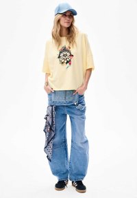Yellow oversized t-shirt with floral graphic, paired with light blue wide-leg denim jeans featuring a scarf detail. Blue cap and sneakers complete the look.
