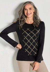 Schwarzes Langarmshirt mit einem Überkreuzmuster aus goldenen Pailletten auf der Vorderseite, kombiniert mit beigen Hosen. Glatte Textur, runder Ausschnitt.