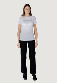 T-shirt bianco in cotone con logo argento "ARMANI EXCHANGE". Abbinato a pantaloni neri e sneakers neri, in piedi davanti a uno sfondo neutro.