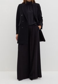 Manteau noir à double boutonnage avec une coupe ajustée, porté sur un haut à col montant et un pantalon à jambes larges, avec des détails à boutons et une texture lisse.