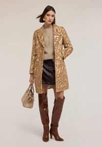 Abrigo de estampado animal marrón sobre un suéter de punto beige, combinado con una falda mini de cuero negra y botas de tacón marrones hasta la rodilla. Incluye un bolso marrón.