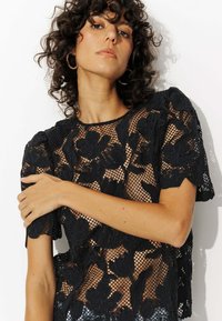 Haut en dentelle noire avec broderie florale, manches courtes bouffantes et design en maille transparente. Encolure ronde et tissu texturé.