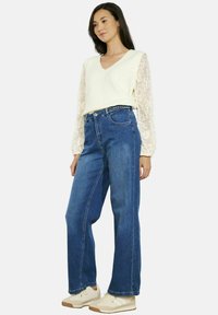 Suéter de cuello en V color crema claro con mangas de encaje, combinado con jeans de mezclilla azul de tiro alto y zapatillas beige, con un corte relajado.