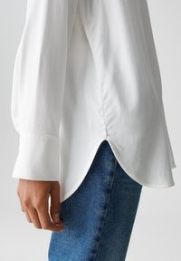 Persoon in een wit overhemd met lange mouwen en een gebogen zoom, over blauwe denim jeans, met een hand ontspannen aan de zijkant.