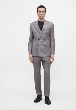 Homme debout portant un costume gris croisé à fines rayures, chaussures noires et chemise noire, sur fond blanc uni.