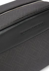 Γωνία μαύρης τσάντας από υφή δέρματος με ασημένιο φερμουάρ και ανάγλυφο λογότυπο Tommy Hilfiger σε λείο μαύρο λουρί δέρματος.