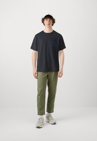 Μαύρο βαμβακερό T-shirt με μπλε εκτυπωμένη λεπτομέρεια στην τσέπη, συνδυασμένο με χακί tapered παντελόνι και μπεζ αθλητικά παπούτσια. Απλή, casual εμφάνιση.