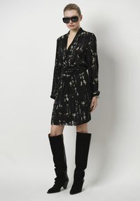 Zwarte jurk met een losse fit, lange mouwen en een ceintuur in de taille; heeft een groen abstract patroon. Gepaard met hoge zwarte laarzen.