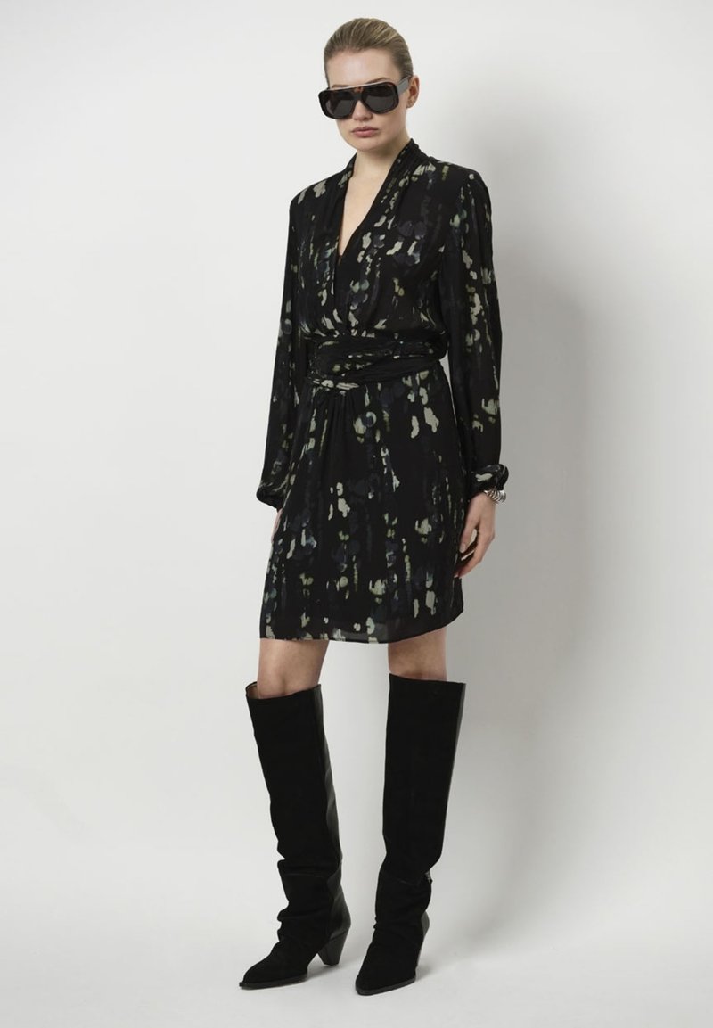 Zwarte jurk met een losse fit, lange mouwen en een ceintuur in de taille; heeft een groen abstract patroon. Gepaard met hoge zwarte laarzen.